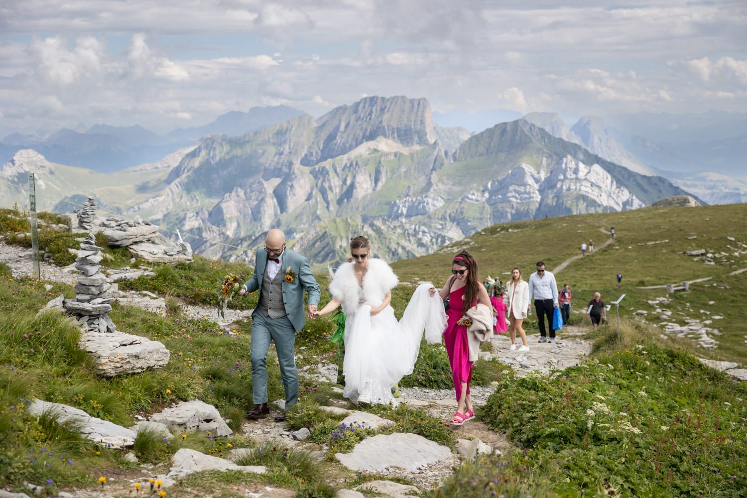 Das Brautpaar läuft Hand in Hand über den Bergpfad Richtung Bergrestaurant, umgeben von Gästen – Hochzeit Chäserrugg.