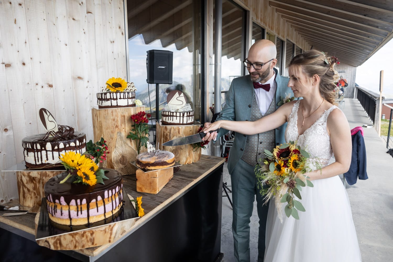 Das Brautpaar schneidet gemeinsam die Hochzeitstorte an – dekoriert mit Sonnenblumen, fotografiert auf dem Chäserrugg.